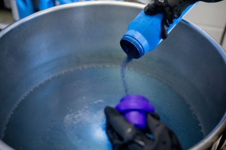Close up of hands, wearing rubber gloves, pouring liquid into a large pot.
