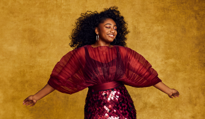 Samara joy spreading her arms wearing a red dress with large sleeves. 